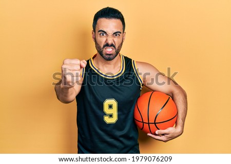 Handsome hispanic man with beard holding basketball ball annoyed and frustrated shouting with anger, yelling crazy with anger and hand raised 
