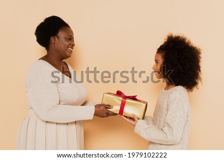 side view of smiling african american granddaughter and grandmother holding gift box on beige background Royalty-Free Stock Photo #1979102222