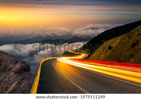 Fast car headlights on European highways. Cargo transportation theme on the highway at sunset. View of traffic and highway running through the mountains. car lights on the highway in the mountain. Royalty-Free Stock Photo #1979111249