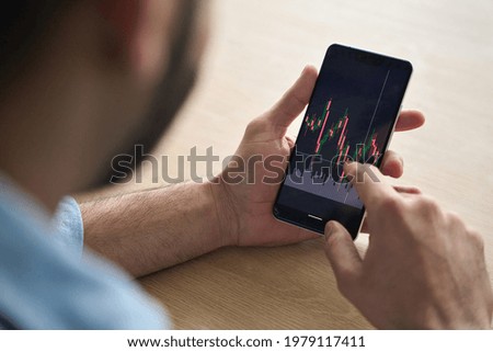 Over shoulder close up view of male analyst broker's hands holding touch screen device smartphone using tech ecommerce application of finance stock markets with graphs and numbers. Royalty-Free Stock Photo #1979117411
