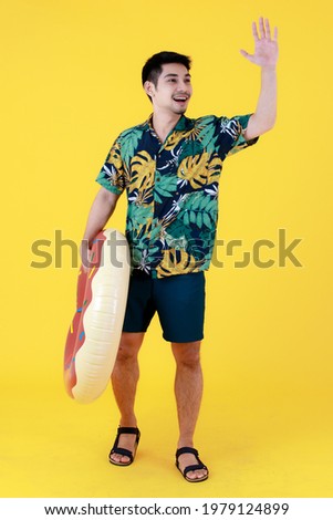 Young Asian man in colorful Hawaiian shirt cheerfully waves hand to greet people while holding swim ring. Full body studio portrait on yellow background. Summer holiday travel concept