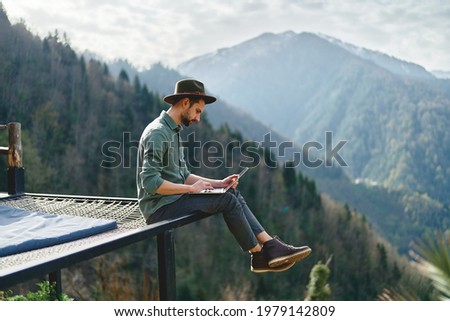 Young man freelancer traveler wearing hat anywhere working online using laptop and enjoying mountains view Royalty-Free Stock Photo #1979142809