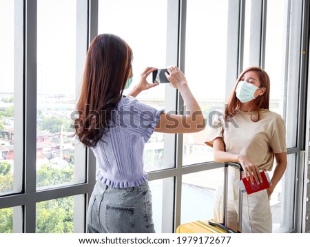 Asian young tourist women wearing face mask to prevent coronavirus infection using mobile phone for taking photos during waiting at airport terminal, social distancing and new normal travel concept.