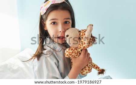 Portrait of a little girl wearing pajama in stripes blue  
and white, smiling broadly, playing with toy giraffe in the bed, isolated on light blue background. Kid has joyful expression in the morning.