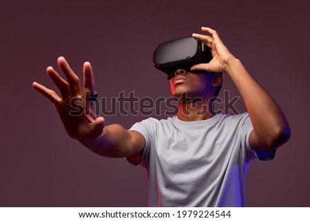 Portrait of young african teen boy in VR helmet looks away at copy space, isolated in studio, guy holds out hand forward. Gamer playing VR games on colourful background, VR gaming.