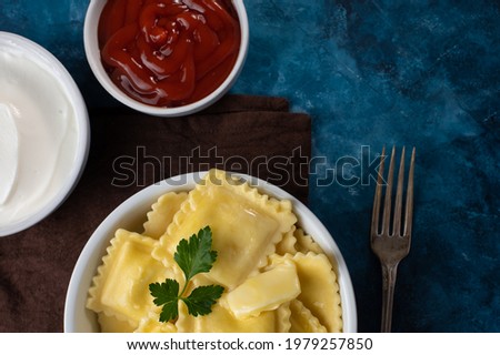 Pictured is a white plate with ravioli. Top decoration is a sprig of mint. there is a fork nearby. Also in the frame are tomato sauce and sour cream. Brown napkin, blue tone.