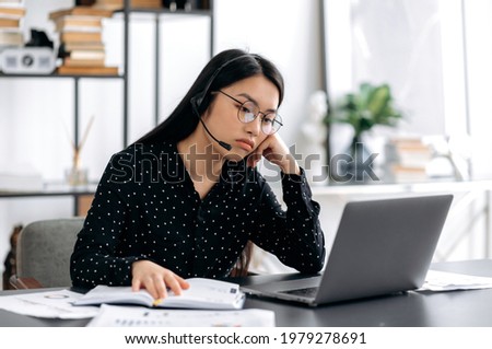 Upset young beautiful asian woman in glasses and headset, call center worker, manager or freelancer, sitting at table in the office, looking sadly at the laptop screen, resting her head on her hand