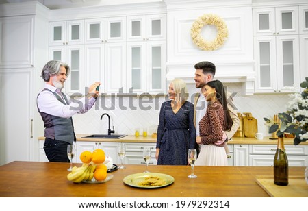 Elegant senior grey haired man grandfather taking picture of his wife and young couple during birthday party taking place in stylish kitchen at home with fruits, sweets and champagne on table