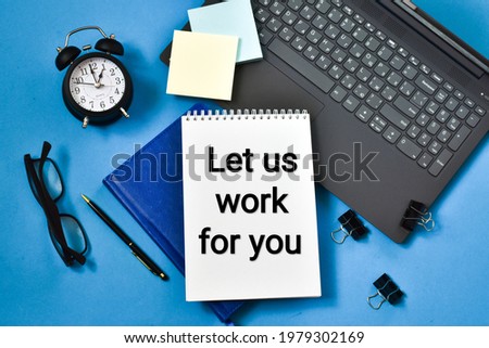 LET US WORK FOR YOU text written in a notepad near a alarm clock, laptop, pen and a eyeglasses on a blue table. Business concept. Flat lay.