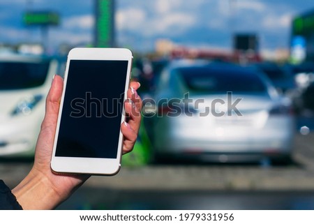 Girl holds a smartphone in his hands. Mock up phone with white screen against the background of an electric car at a charging station