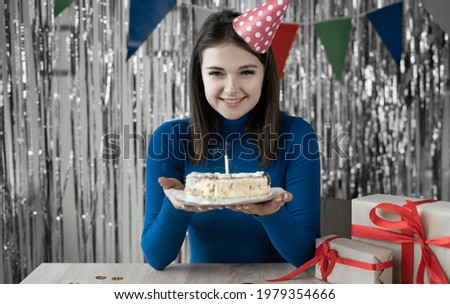 Portrait of a millennial girl sitting at a table with a birthday cake in her hands and a blown out candle. A young lady celebrates one birthday with her friends online on a laptop webcam. House party
