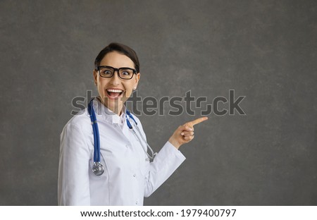 Funny crazy emotional female doctor standing on gray background smiling with wide open mouth and eyes. Woman in medical gown looking at camera pointing finger at free space for text. Banner.