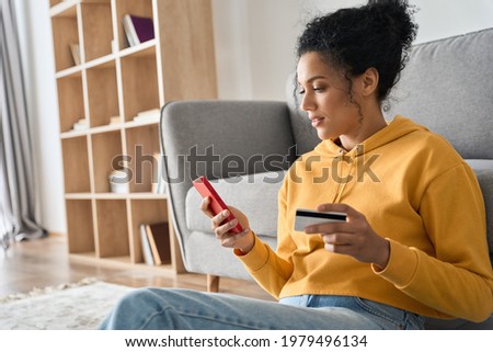 Young adult African American female consumer holding credit card and smartphone sitting on floor at home doing online banking transaction. E commerce virtual shopping, secure mobile banking concept. Royalty-Free Stock Photo #1979496134