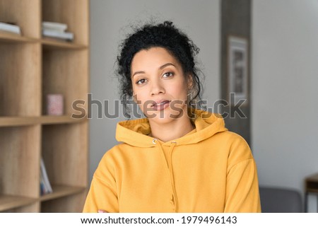 Headshot portrait of young attractive African American mixed race girl student in casual clothing hoodie posing indoors at modern living room home office looking at camera. Portrait Royalty-Free Stock Photo #1979496143