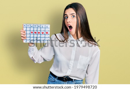 Young brunette teenager holding heart calendar scared and amazed with open mouth for surprise, disbelief face 