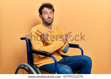 Handsome man with beard sitting on wheelchair pointing to both sides with fingers, different direction disagree 