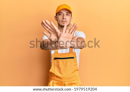 Hispanic young man wearing handyman uniform rejection expression crossing arms doing negative sign, angry face 