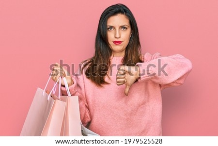 Beautiful hispanic woman holding shopping bags with angry face, negative sign showing dislike with thumbs down, rejection concept 