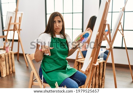 Young hispanic artist women painting on canvas at art studio pointing down looking sad and upset, indicating direction with fingers, unhappy and depressed. 