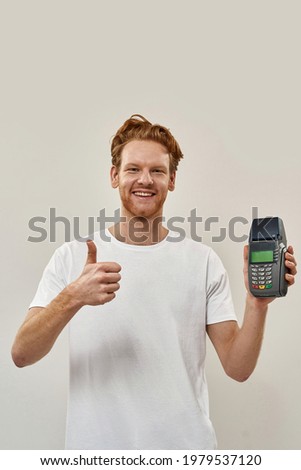 Picture of a smiling young guy with payment terminal in hand and big thumb up, watches straight in camera , wears white tee , stay at home concept