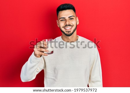 Young hispanic man with beard holding glass of milk looking positive and happy standing and smiling with a confident smile showing teeth 