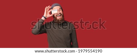 Photo of cheerful bearded hipster man holding debit credit card over eyes and smiling.