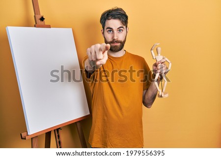 Caucasian man with beard standing by painter easel stand holding manikin pointing with finger to the camera and to you, confident gesture looking serious 