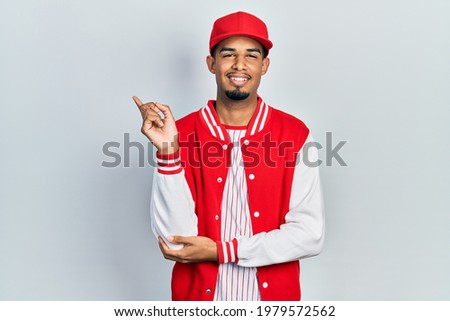 Young african american man wearing baseball uniform with a big smile on face, pointing with hand and finger to the side looking at the camera. 