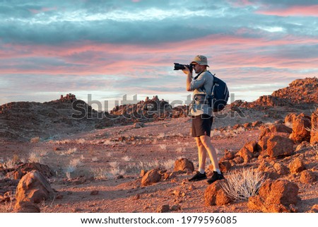 Photographer taking photo in rocks of Namib Desert, Namibia, Africa. Red mountains and sunset sky in background. Landscape photography