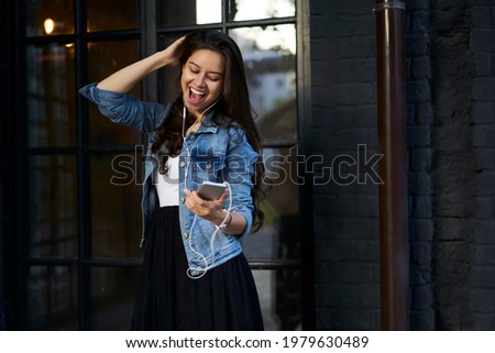 Cheerful female blogger browsing content in social media using 4g wireless on mobile phone, excited hipster girl with smartphone technology enjoying time for listening positive music playlist