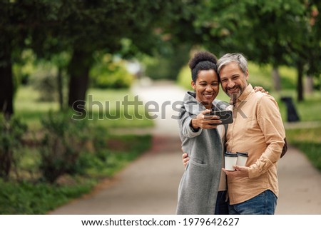 Mature friends, taking a photo in the park, holding cups of coffee.