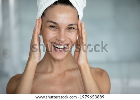Head shot of happy woman with head wrapped in bath towel, touching face with toothy smile, applying cream, oil, lotion on facial skin after shower in bathroom. Skincare therapy concept. Portrait