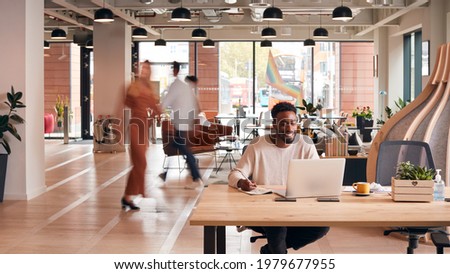 Businessman Sitting At Desk Writing In Notebook In Modern Open Plan Office