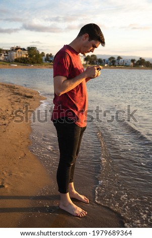 A man taking a photo of his feet on the beach barefoot. He is looking at his mobile phone while taking the picture. It is getting dark.