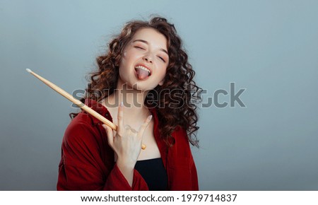 Rock and roll music concept. Cute caucasian young woman shows her tongue and and holds drumsticks in his hands on a gray-blue studio background. Free space for advertisement.