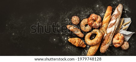 Bakery - various kinds of breadstuff. Bread rolls, baguette, bagel, sweet bun and croissant captured from above (top view, flat lay). Black background, free copy space. Horizontal banner layout. Royalty-Free Stock Photo #1979748128