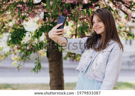 Young smiling woman taking selfie self portrait photos on smartphone. Model posing on park sakura trees background. Female showing positive face emotions.