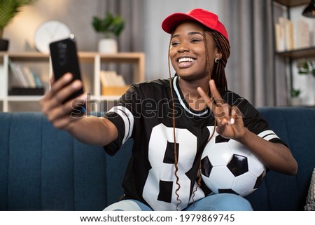 Pretty african woman holding soccer ball while sitting on sofa and and taking selfie on modern smartphone. Happy football fan watching world cup on TV from home.