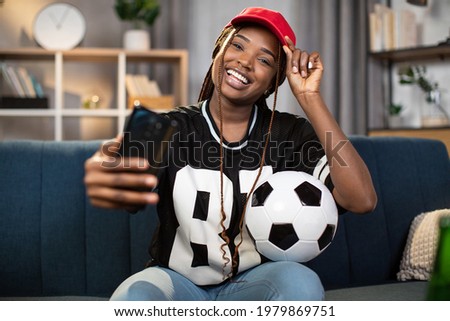 Happy black woman using modern smartphone for taking selfie while sitting on comfy couch at home. Smiling female staying at home for soccer match on TV