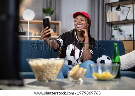 Smiling african american woman taking selfie on modern smartphone while watching soccer game at home. Concept of people and technology.