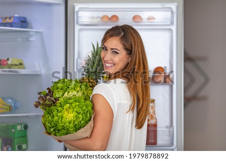 Happy young woman standing at the opened fridge. Portrait of cute serious female standing near open fridge full of healthy food, vegetables and fruits, healthy lifestyle concept