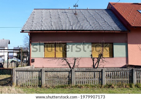 Old small attached suburban family house in need of restoration with faded facade and closed dilapidated window blinds surrounded with concrete fence and uncut grass on clear blue sky background