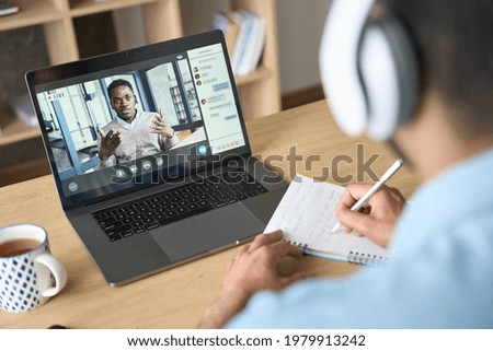 Latin indian adult student wearing headset having virtual meeting online call training educational webinar chatting with black teacher at home office using laptop. Over shoulder view. Royalty-Free Stock Photo #1979913242