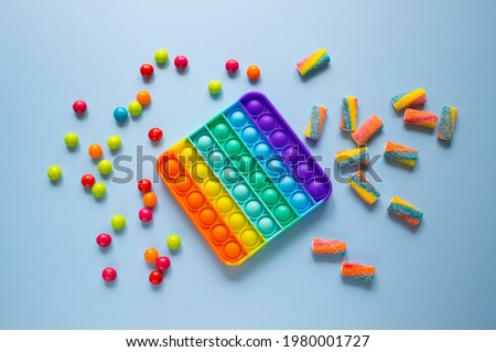 Rainbow Pop it fidget toy next to the colorful dragees and marmalade. Poppit - new fidget antistress toy for children on blue background. Royalty-Free Stock Photo #1980001727