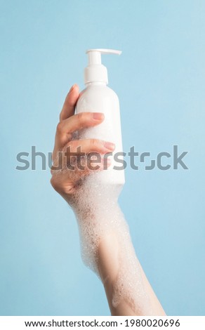 Womans hand holding cosmetics bottle with foam bubbles on pastel blue background. Bath accessories for body care, spa beauty treatment and skincare concept. Vertical, close up Royalty-Free Stock Photo #1980020696