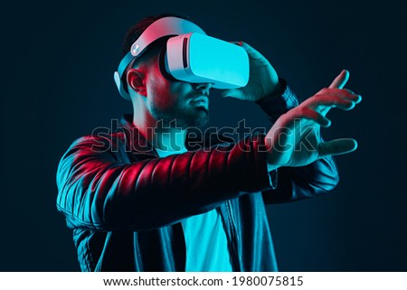 Bearded guy in black leather jacket and VR goggles interacting with invisible screen while experiencing virtual reality in dark studio with neon lights