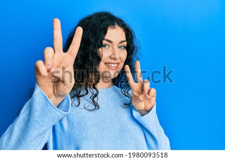 Young brunette woman with curly hair wearing casual clothes smiling looking to the camera showing fingers doing victory sign. number two. 