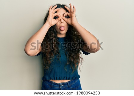 Young brunette woman with curly hair wearing casual clothes doing ok gesture like binoculars sticking tongue out, eyes looking through fingers. crazy expression. 