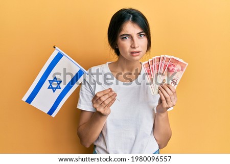 Young caucasian woman holding israel flag and shekels banknotes clueless and confused expression. doubt concept. 
