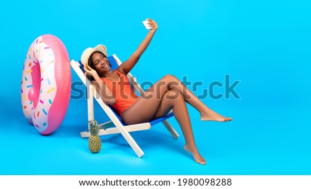 Gorgeous black woman taking selfie with smartphone in lounge chair over blue background, banner with free space. Positive young lady making photo of herself in bikini, having cool summer vacation